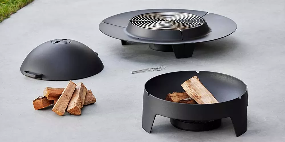 Cane-line Ember Kollektion in Schwarz, bestehend aus Feuerschale und Grillaufsatz, auf grauem Betonboden im Freien präsentiert.