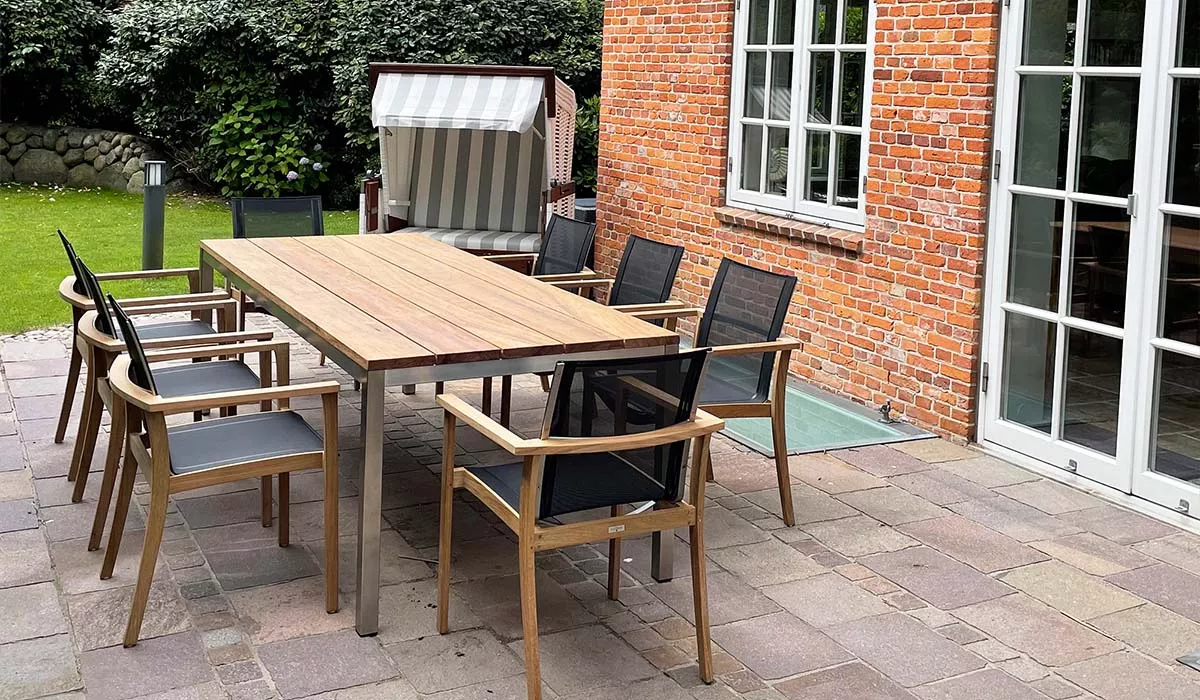 Terrasse mit Royal Mirage Sitzmöbeln in Holzoptik und schwarzen Polstern, umgeben von Backsteinmauer und Garten im Hintergrund.