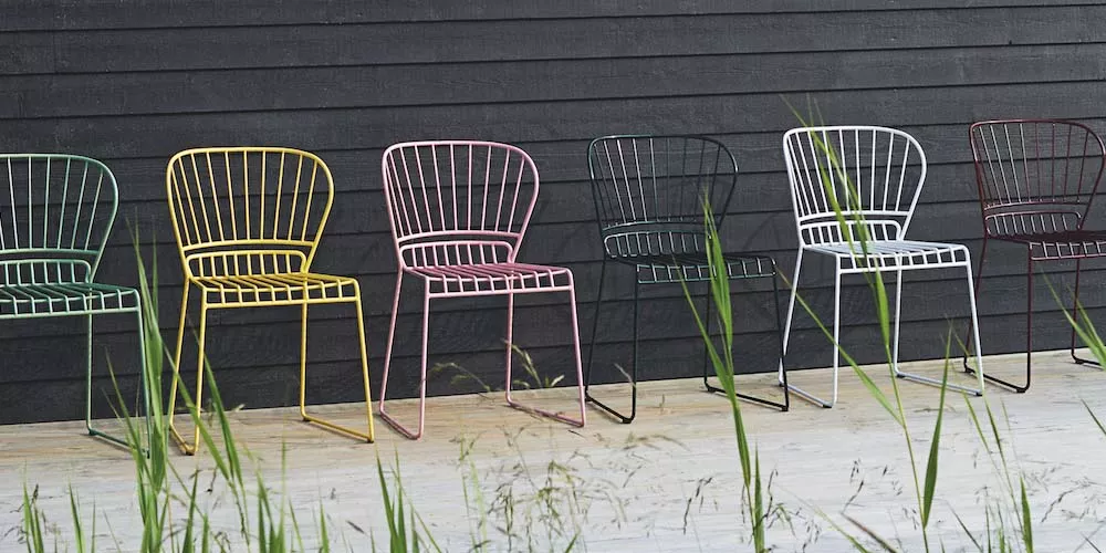 Sechs Skargaarden Resö Stühle in verschiedenen Farben stehen auf einer Holzterrasse vor einer dunklen Holzwand, umgeben von Gräsern.