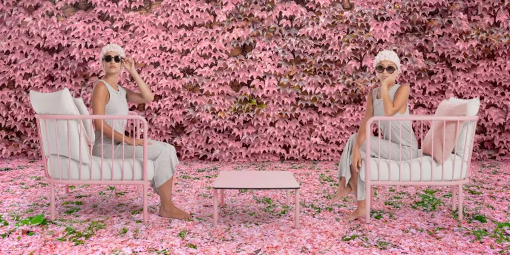 Zwei Personen in grauen Outfits sitzen auf rosa Diabla Arp Stühlen vor einer Wand aus rosa Blättern, mit einem passenden Tisch dazwischen.