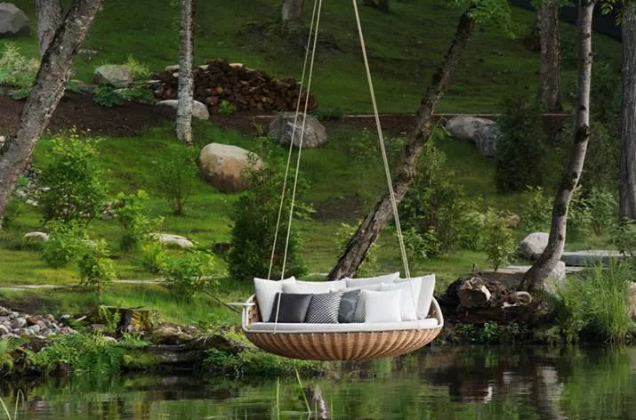Dedon Swingrest Hanging Lounger hängt an Seilen über einem ruhigen Teich in einem grünen Waldumfeld mit Kissen dekoriert Mood Bild