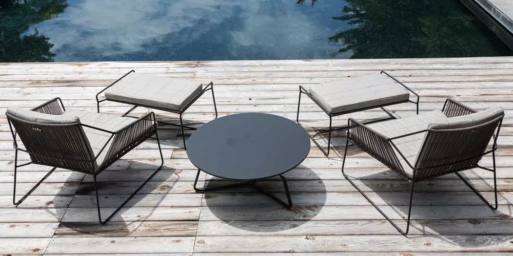 Oasiq Sandur Loungegruppe in Grau auf Holzterrasse am Pool, bestehend aus zwei Sesseln, zwei Hockern und einem runden Tisch.