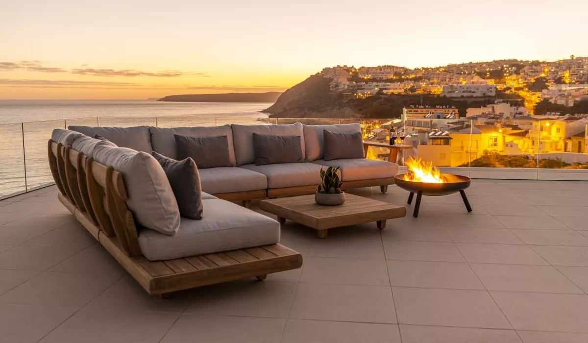 Alexander Rose Lounge-Sofa in Holzoptik mit grauen Kissen auf einer Terrasse bei Sonnenuntergang, mit Blick auf das Meer und die Stadt.