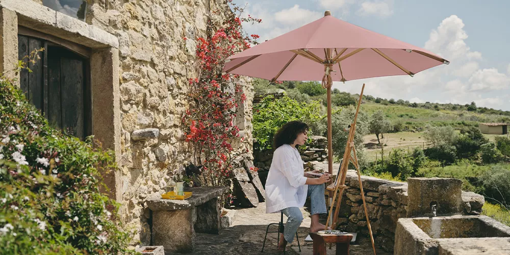 Frau sitzt unter einem rosa Glatz Teakwood Sonnenschirm auf einer rustikalen Terrasse mit Steinmauer und blühenden Pflanzen im Hintergrund.