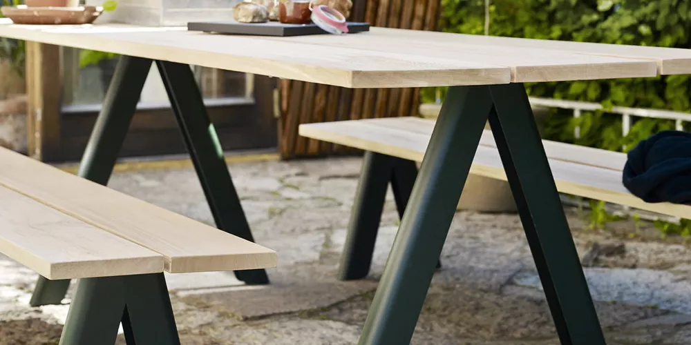 Skagerak Overlap Tisch und Bänke in hellem Holz und dunklem Metall auf einer Terrasse mit grünem Hintergrund.