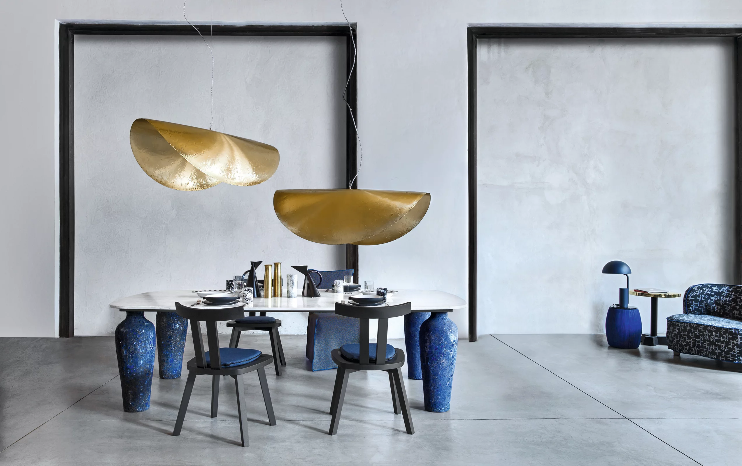 Zwei goldene Foscarini Hängeleuchten über einem modernen Esstisch mit blauen Stühlen in einem minimalistischen Raum.