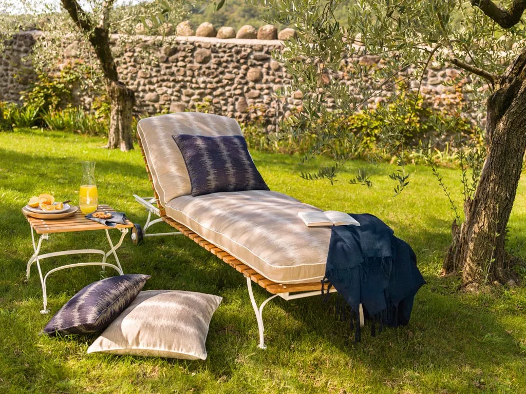 Sonnenliege Classic von Weish in Beige auf grünem Rasen mit Kissen und Beistelltisch, umgeben von Natursteinmauer und Bäumen.