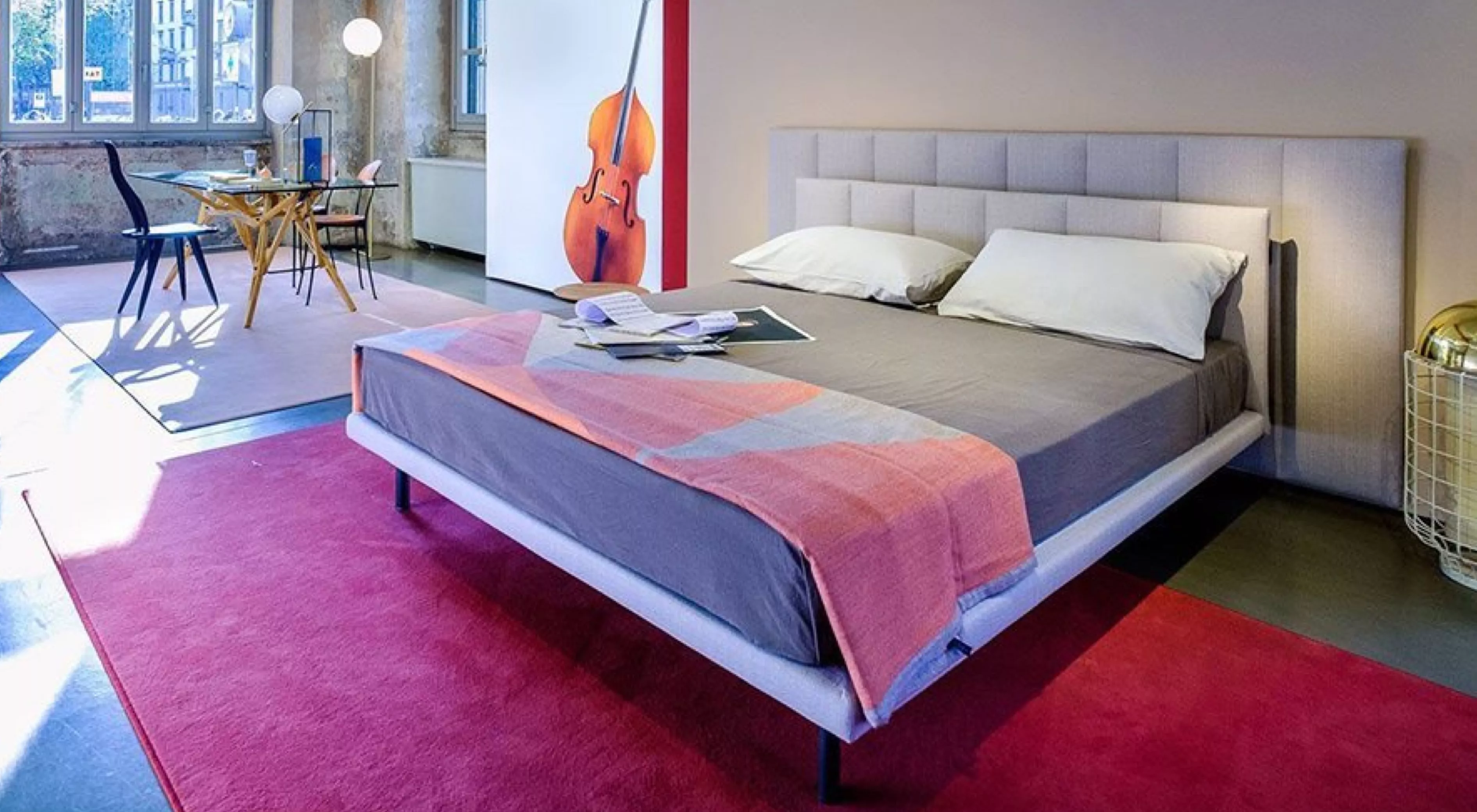 Ein modernes Schlafzimmer mit einem hellgrauen Polsterbett auf einem roten Teppich, im Hintergrund ein Tisch und große Fenster.