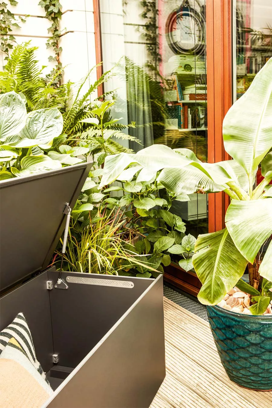 Conmoto El Pecho Kissentruhe in Schwarz, 180x90 cm, steht auf einer Terrasse umgeben von üppigen Pflanzen vor einer Glasfassade.