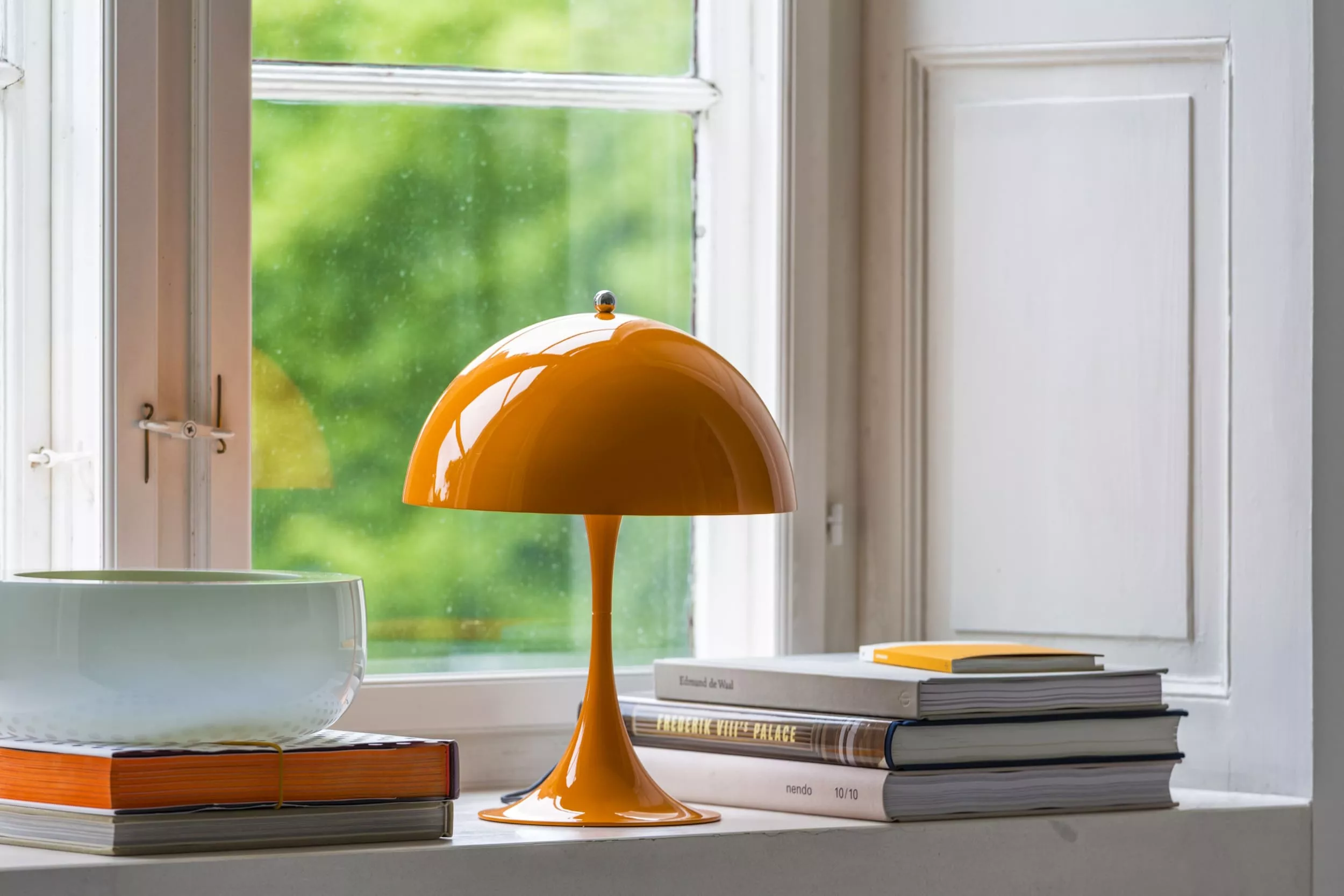 Orangefarbene Louis Poulsen Panthella 250 Tischlampe auf Fensterbank mit Büchern und Schale, vor grünem Hintergrund.
