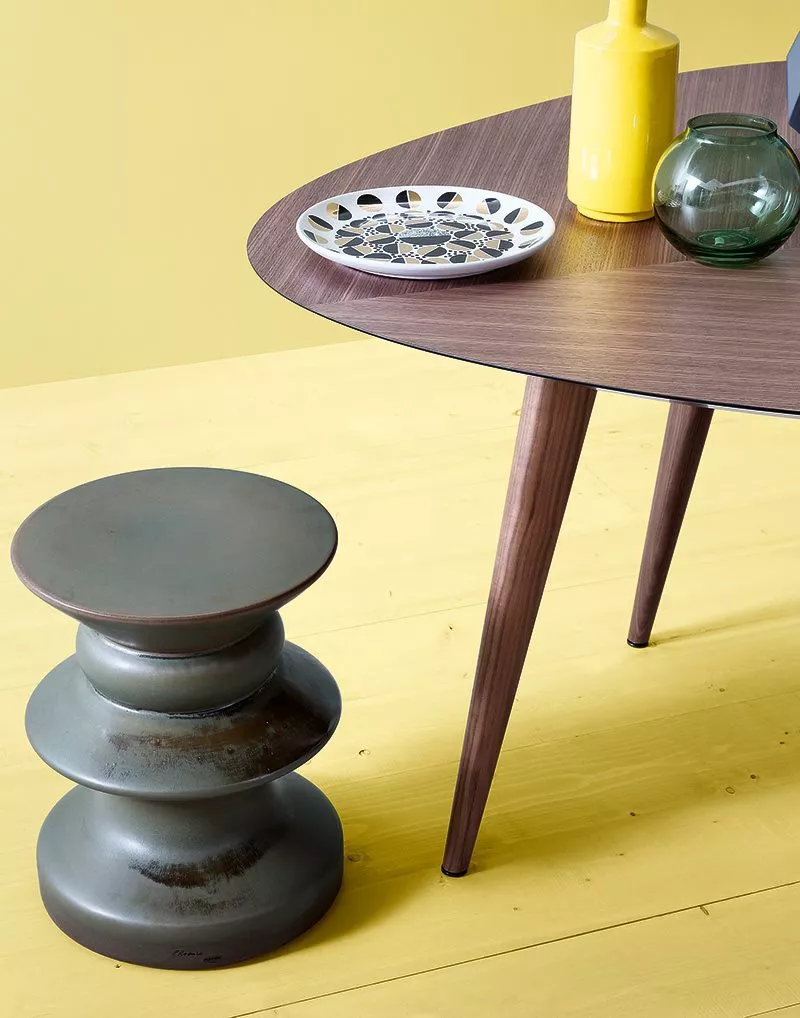 Ein Zanotta Teti Hocker steht neben einem runden Holztisch auf einem gelben Boden mit dekorativen Vasen und einer Schale darauf