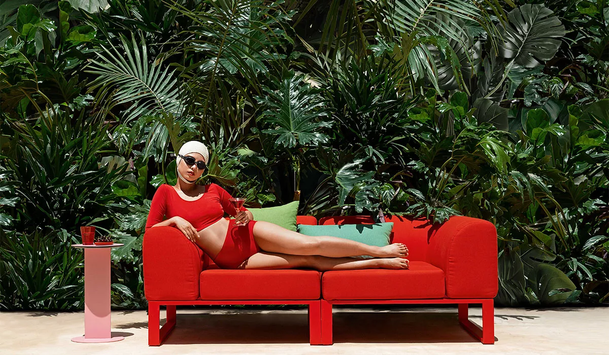 Frau in rotem Badeanzug auf rotem Diabla Loungemöbel, umgeben von tropischen Pflanzen, mit rosa Beistelltisch und Getränk.