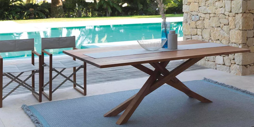 Talenti Bridge Gartenmöbel-Kollektion in Holzoptik auf einer Terrasse mit Pool im Hintergrund, zeigt Tisch und Stühle in natürlicher Umgebung.