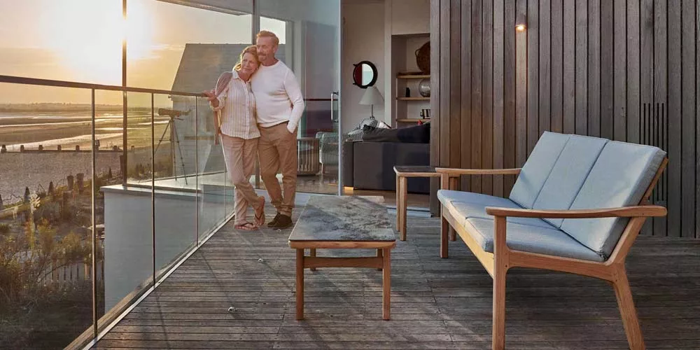 Barlow Tyrie Monterey Loungegruppe in hellem Holz auf einer Terrasse mit Meerblick bei Sonnenuntergang, gemütliche Stimmung.