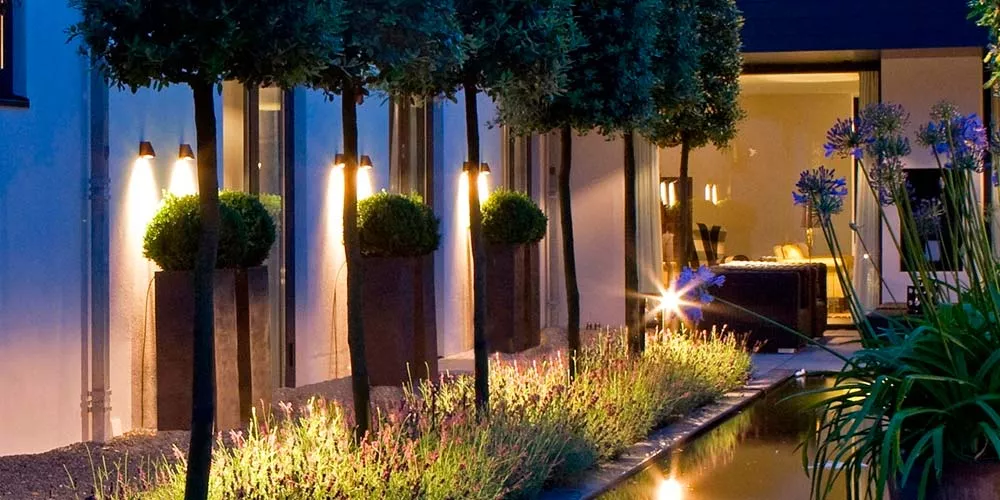 Royal Botania Beamy Gartenleuchten in warmem Licht, beleuchten stilvoll einen modernen Garten bei Nacht, umgeben von Pflanzen und Wasser.