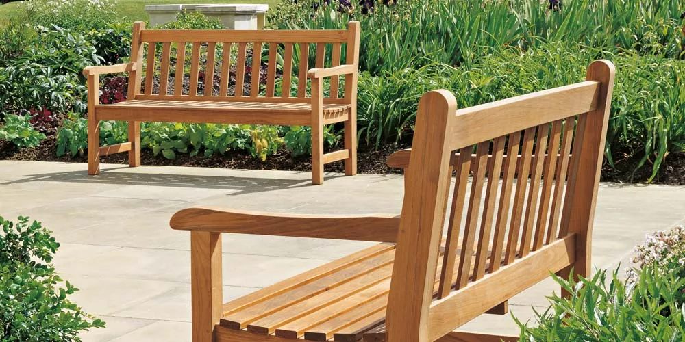 Zwei Barlow Tyrie Felsted Holzbänke in natürlicher Umgebung auf einer Terrasse, umgeben von grünen Pflanzen.