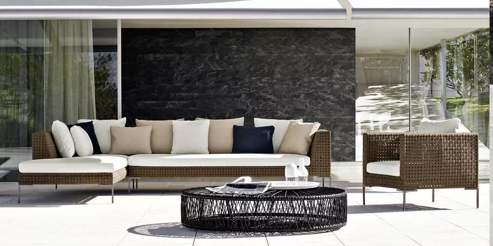 BeB Italia Charles Outdoor Loungegruppe mit Loungesofa und Sessel auf einer Terrasse vor einer modernen Glasfassade und einem dunklen Steinwandhintergrund