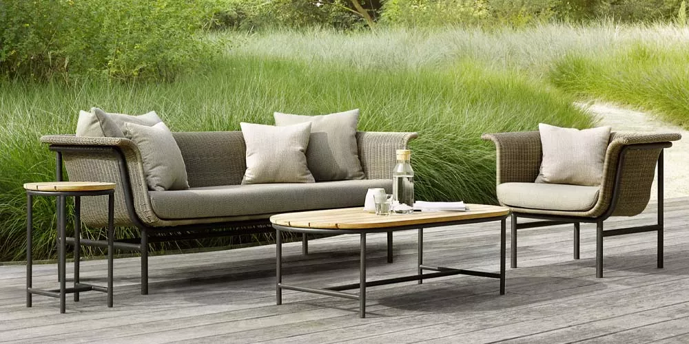 Vincent Sheppard Wicked Gartenmöbel-Set in Naturtönen auf Holzterrasse vor grünem Hintergrund, bestehend aus Sofa, Sessel und Tisch.