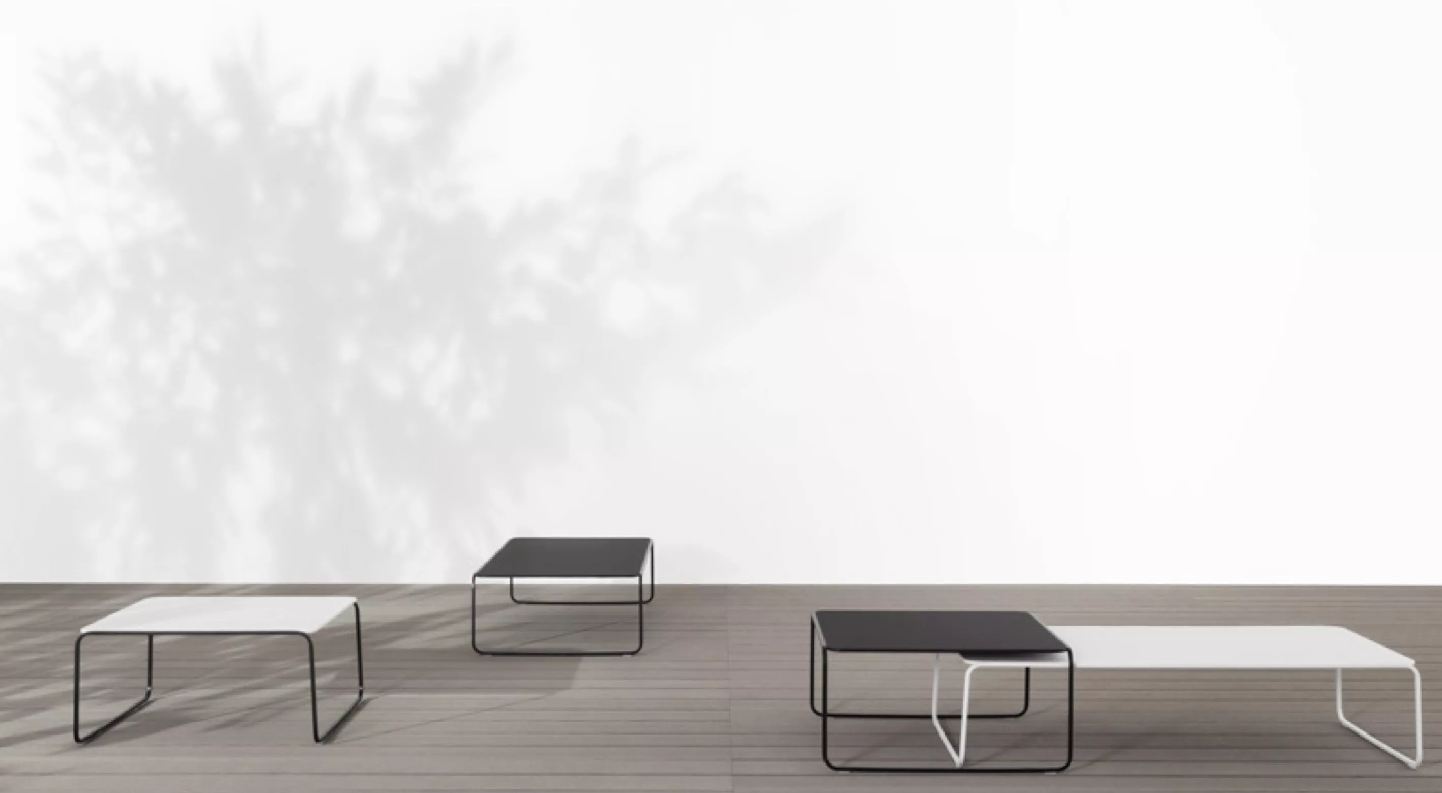 Drei TOOU TO-E Outdoor-Tische in Schwarz und Weiß auf einer Holzterrasse vor einer weißen Wand mit Schatten.