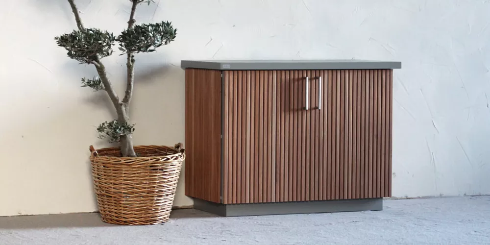 Cubic Outdoor Living Box in Holzoptik, braun, neben einem Korb mit Pflanze vor einer weißen Wand auf grauem Boden.