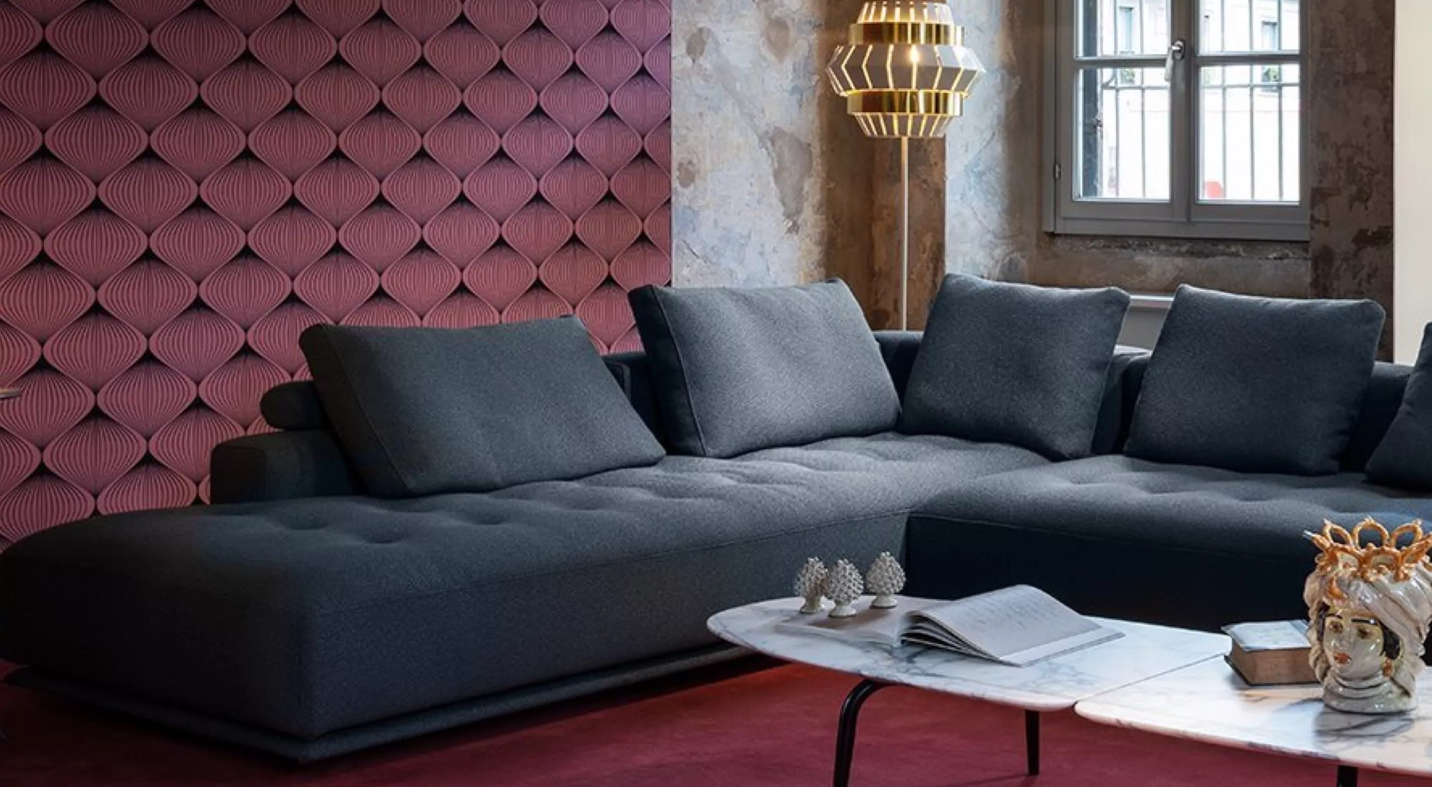 Ein modernes Wohnzimmer mit einem grauen Zanotta Shiki Sofa einem weißen Marmortisch und einer goldenen Lampe vor einer roten Wand.