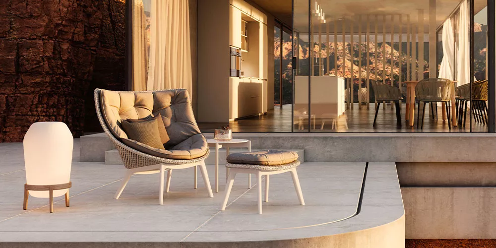 Dedon Loungesofa Mbrace steht auf einer Terrasse vor einem modernen Haus mit Glasfront und bietet eine entspannte Sitzgelegenheit in der Abendsonne