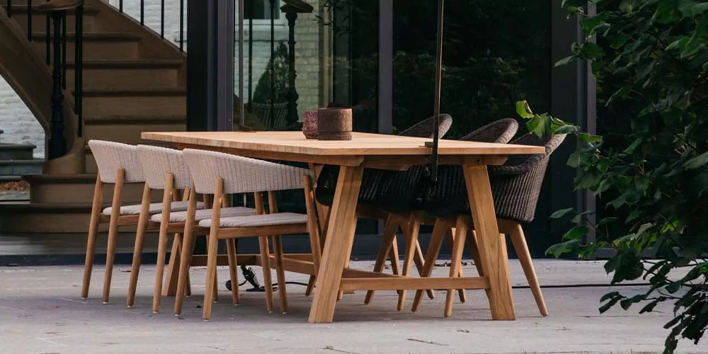 Vincent Sheppard Mona Esstisch-Set aus hellem Holz mit sechs Stühlen auf einer Terrasse vor einem modernen Gebäude.