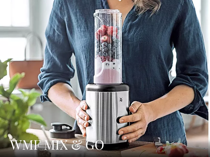 Frau in Küche hält silbernen WMF Mix & Go Smoothiemaker mit frischen Beeren, Hintergrund unscharf.