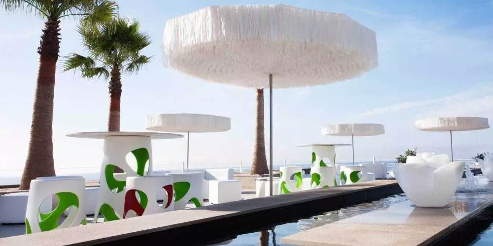 Frou Frou Sonnenschirme in Weiß auf einer stilvollen Terrasse mit Palmen und modernem Mobiliar, Blick auf das Meer im Hintergrund.