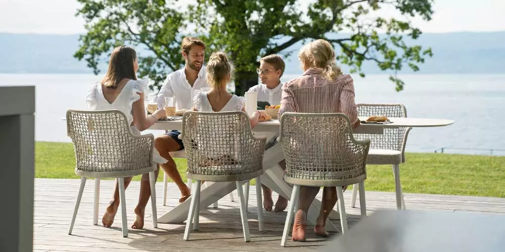 Familie sitzt an einem weißen Varaschin Emma Dining Gartentisch mit grauen Stühlen auf einer Terrasse mit Seeblick.