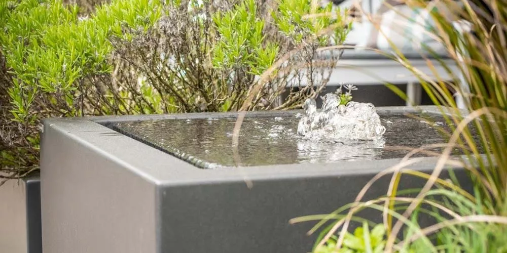 Gartenbrunnen aus grauem Stein mit sprudelndem Wasser, umgeben von grünen Pflanzen, in einem modernen Außenbereich.
