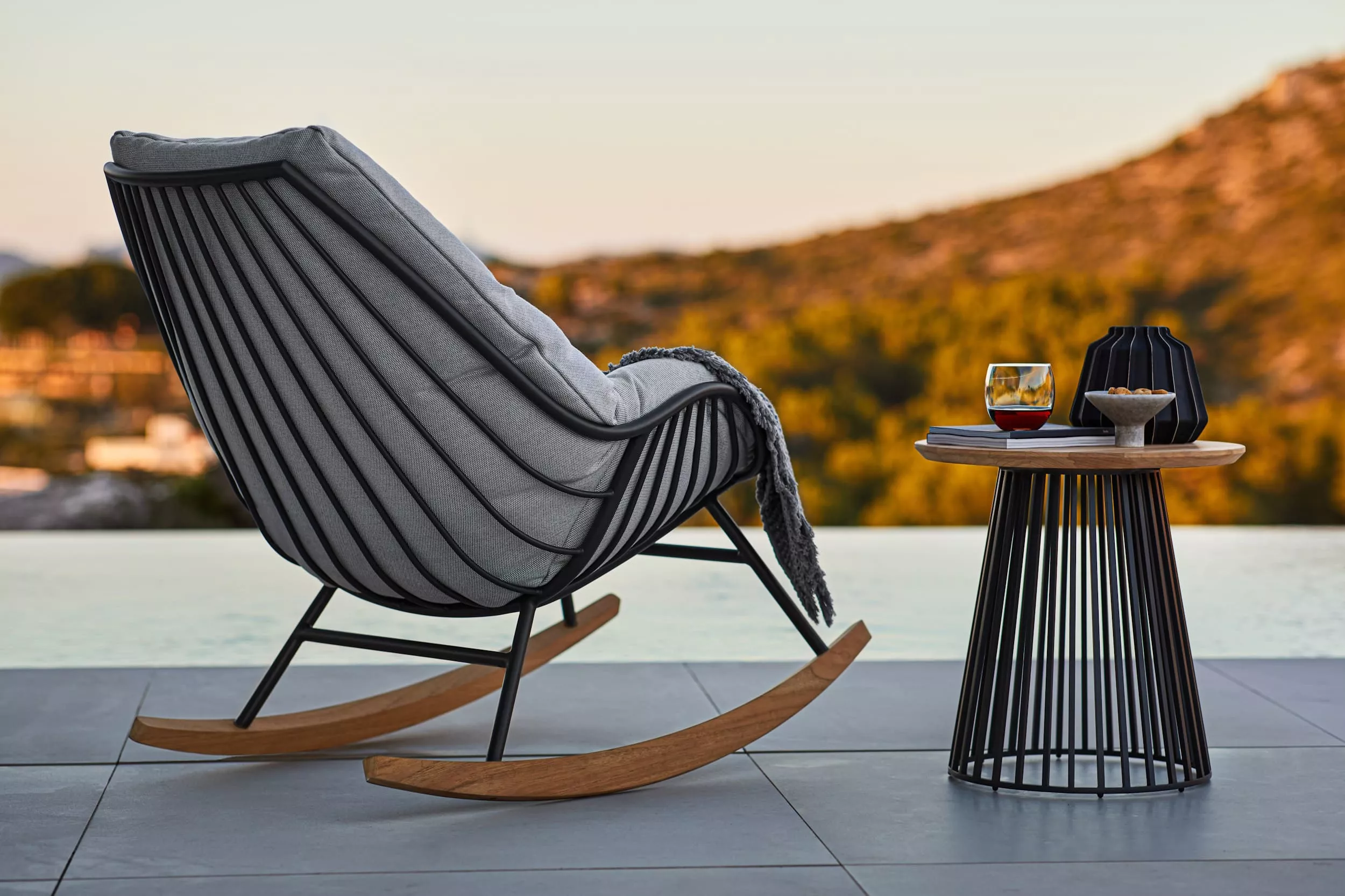 Solpuri Chill-A Schaukelstuhl mit grauem Kissen und Beistelltisch mit Getränk und Snack vor einer Landschaft im Hintergrund Moodbild