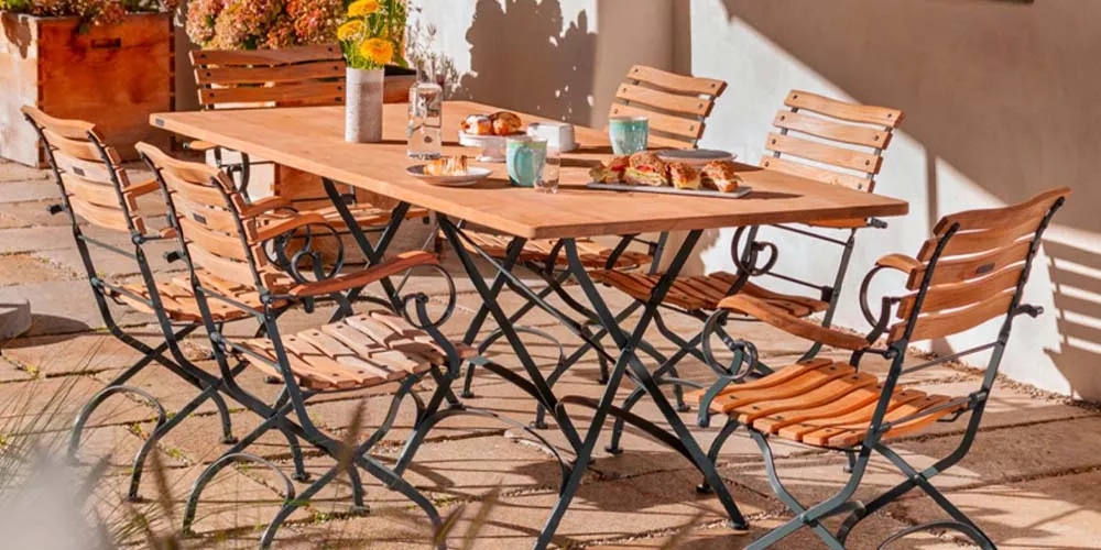 Weißhäupl Classic Gartenmöbel-Set aus Holz und Metall in einem sonnigen Garten mit gedecktem Tisch und Blumen im Hintergrund.