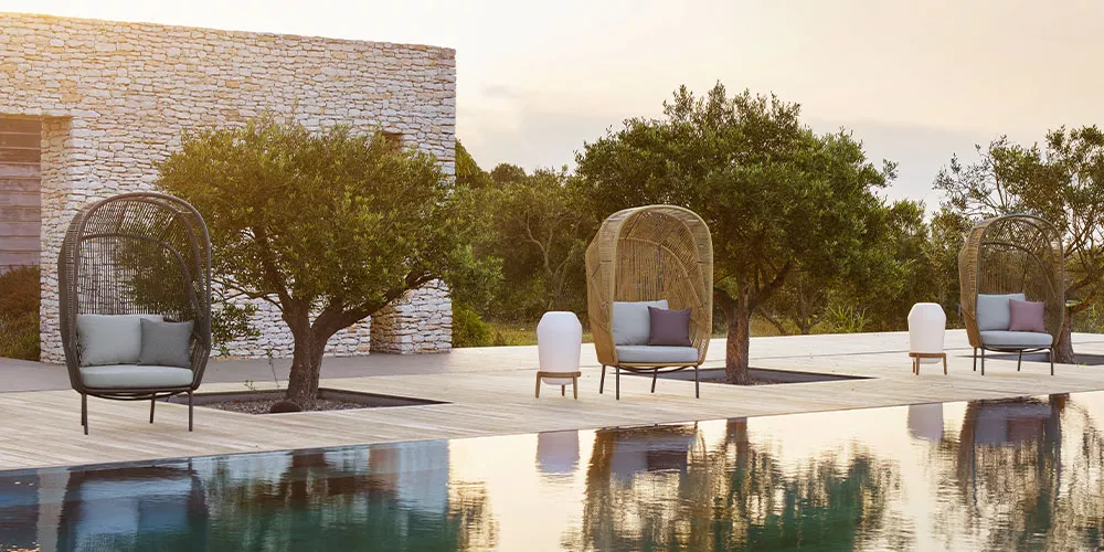 Dedon Rilly Kollektion zeigt stilvolle Outdoor-Sessel und Beistelltische auf einer Terrasse mit Pool und Olivenbäumen im Hintergrund