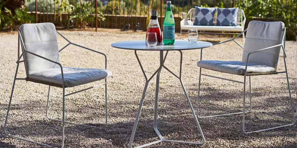 Oasiq Serac Essgruppe in Weiß auf Kiesuntergrund mit zwei Stühlen und rundem Tisch, darauf Wasserflasche und Gläser, im Garten.