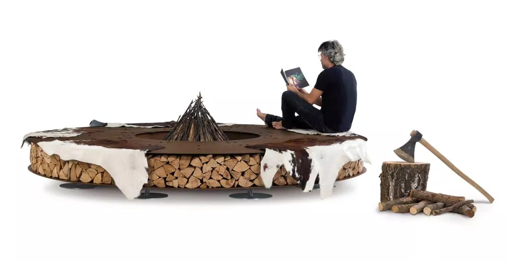 Ein Mann sitzt auf der AK47 Zero Feuerstelle 300 cm Gartenkamin und liest ein Buch daneben liegen Holzscheite und eine Axt auf dem Boden