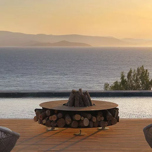 AK47 Zero Feuerstelle 150 cm Gartenkamin auf einer Terrasse mit Blick auf das Meer und die Berge im Hintergrund stimmungsvolles Bild bei Sonnenuntergang