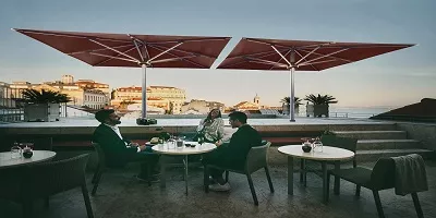 Drei Personen sitzen unter roten Sonnenschirmen auf einer Terrasse mit Blick auf eine Stadtlandschaft bei Sonnenuntergang.