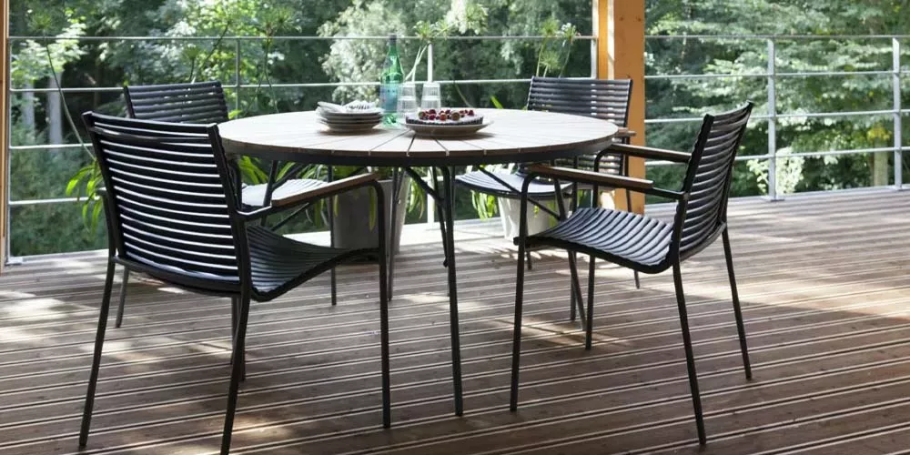 Runder Holztisch mit vier schwarzen Stühlen auf einer Terrasse mit Blick auf grüne Bäume, sommerliche Atmosphäre.