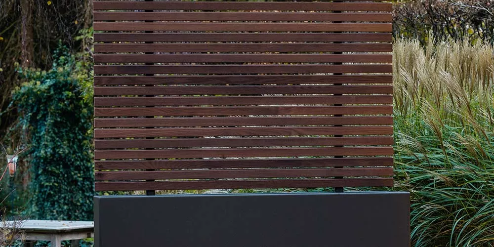 Braunholz-Sichtschutz von Villa Schmidt, 180x180 cm, in einem Garten mit grünem Hintergrund und Gräsern, stimmungsvolle Atmosphäre.