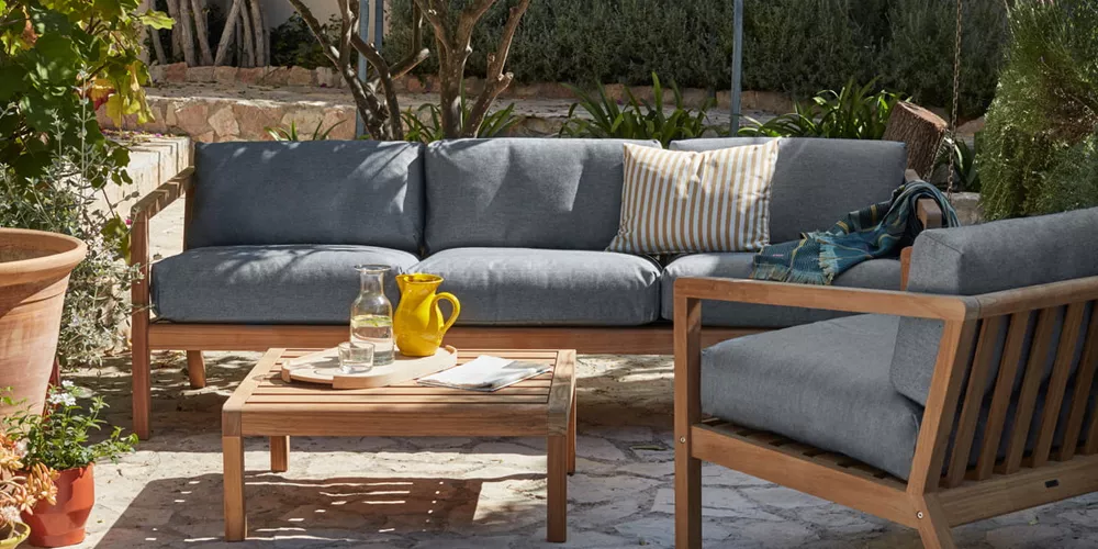 Skagerak Virkelyst Sofa in Grau auf einer Terrasse mit Holztisch, umgeben von Pflanzen, im sommerlichen Ambiente.