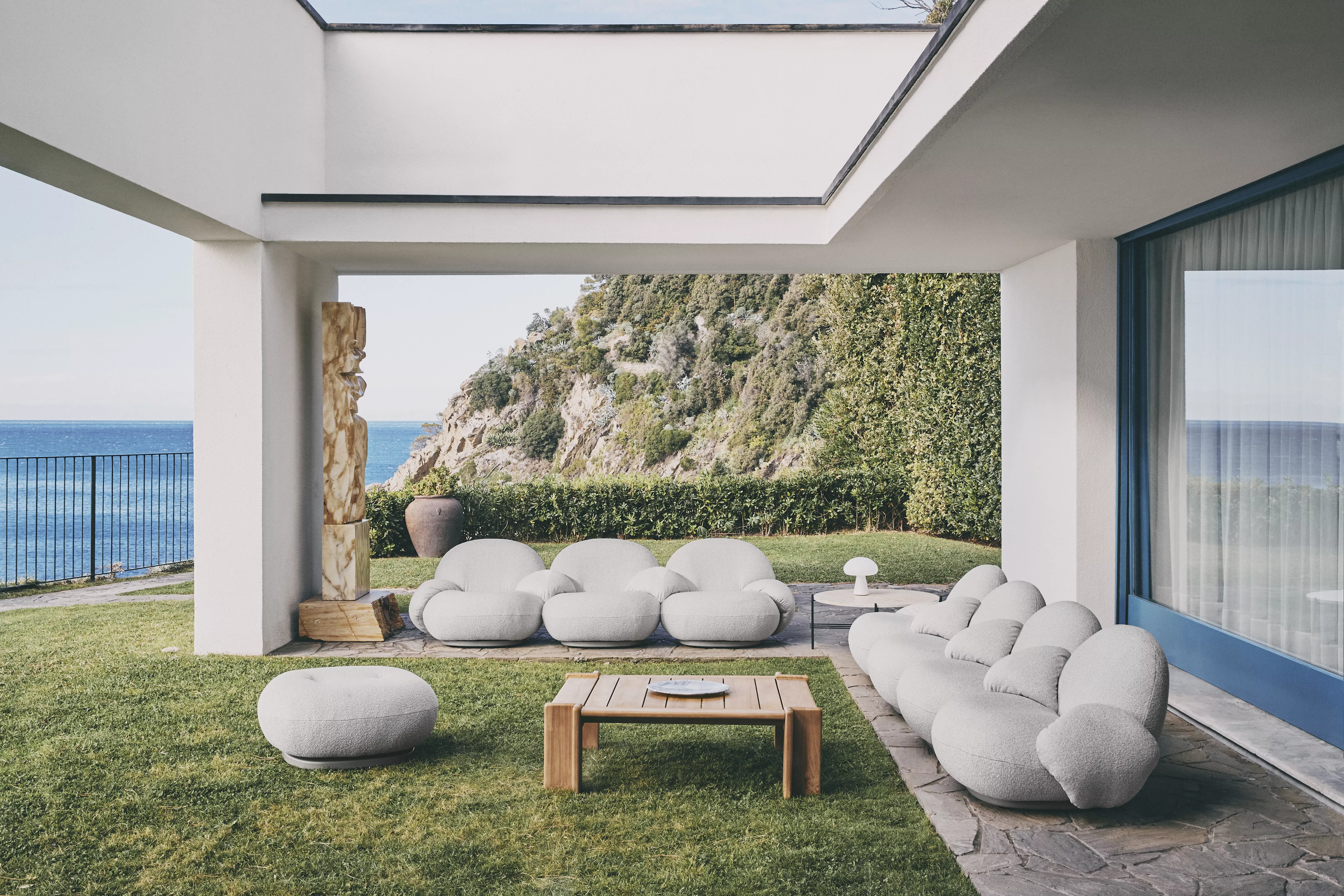 GUBI Pacha Sessel und Obello Lampe auf einer Terrasse mit Meerblick und grüner Landschaft im Hintergrund