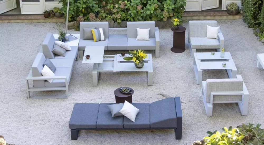 Ego Paris KAMA Gartenmöbel-Set in Grau auf Kiesboden, bestehend aus Sofas, Sesseln und Tischen, dekoriert mit Kissen und Blumen.