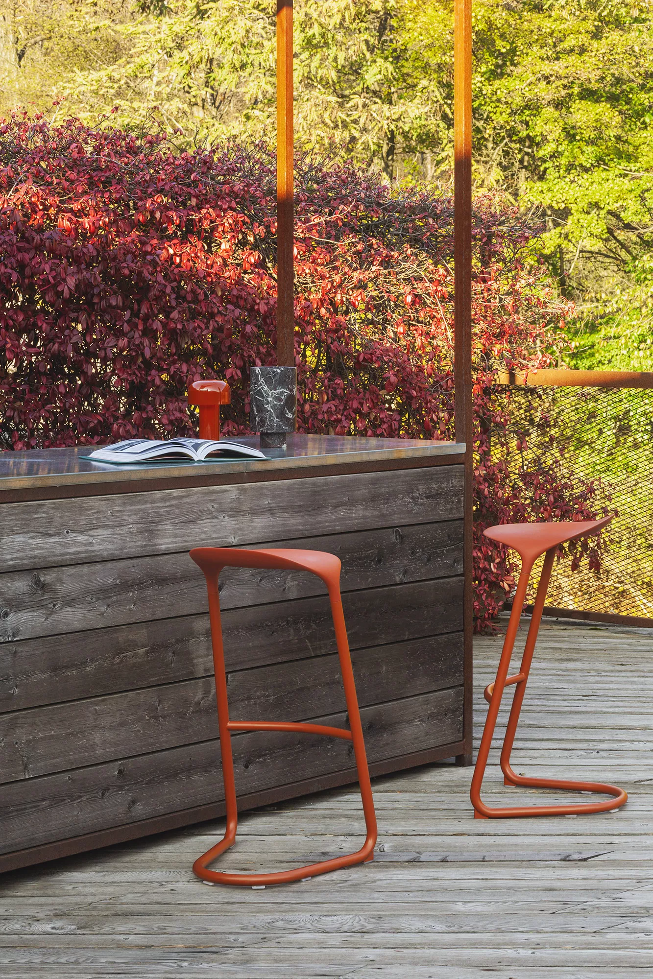 Zwei orangefarbene Vondom Wing Barhocker aus Kunststoff auf einer Holzterrasse vor einer Holzwand, stimmungsvolle Außenaufnahme.