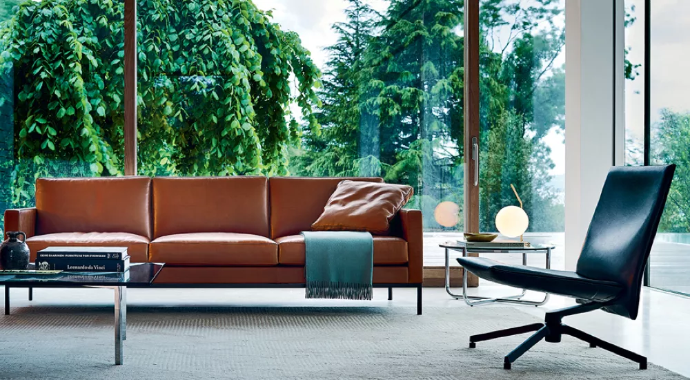 Braunes Ledersofa von Villa Schmidt vor großen Fenstern mit Blick auf grüne Bäume, daneben ein schwarzer Sessel und ein runder Tisch.