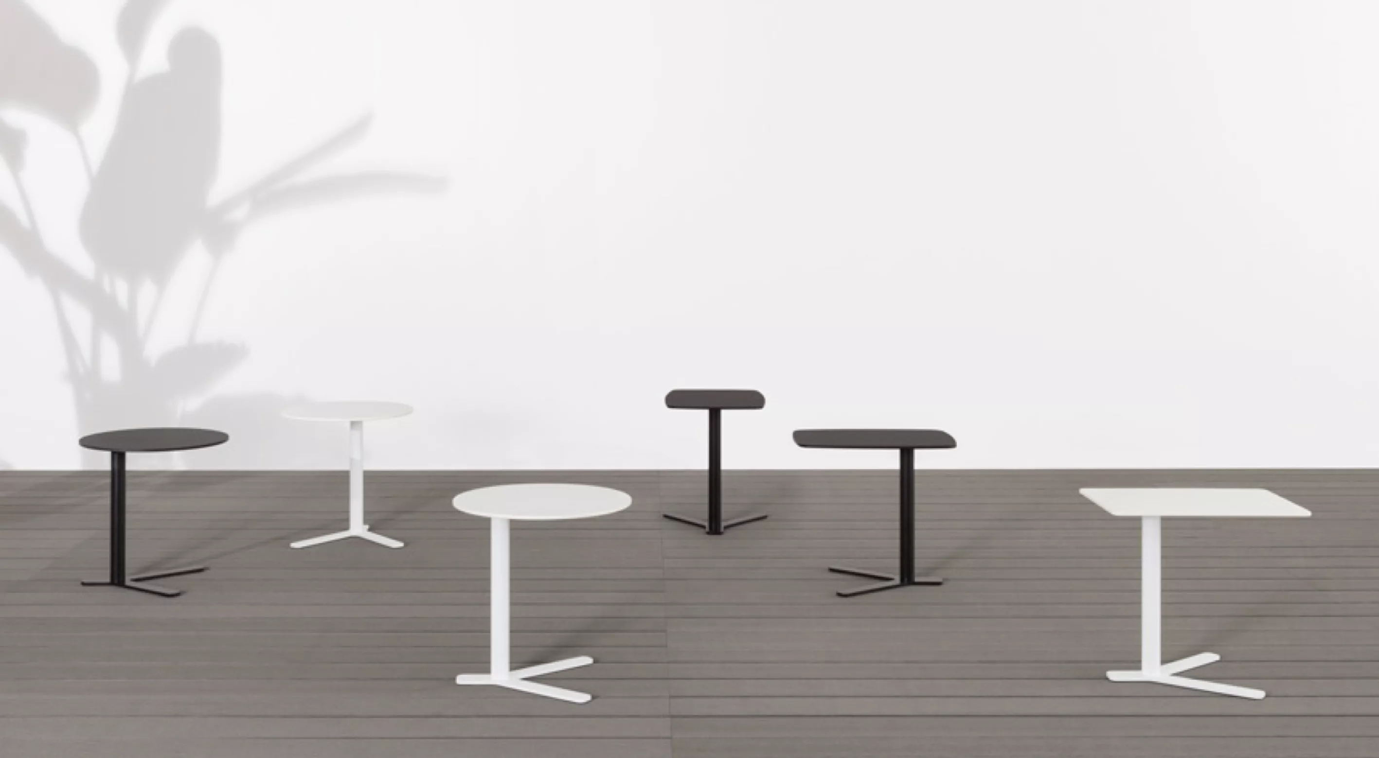 Sechs Yo Outdoor Beistelltische von Kristalia in Schwarz und Weiß auf grauem Holzboden vor einer weißen Wand mit Pflanzenschatten.