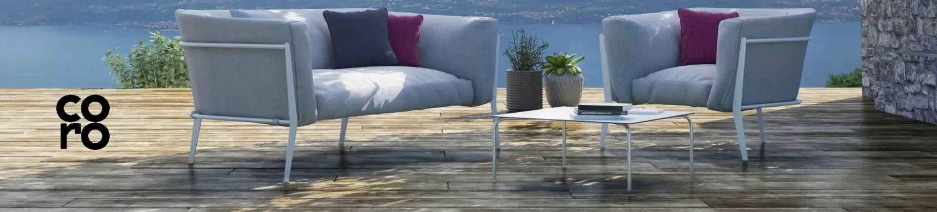 Zwei graue CORO-Sessel und ein weißer Tisch auf einer Holzterrasse mit Seeblick, dekoriert mit Pflanzen und Kissen.