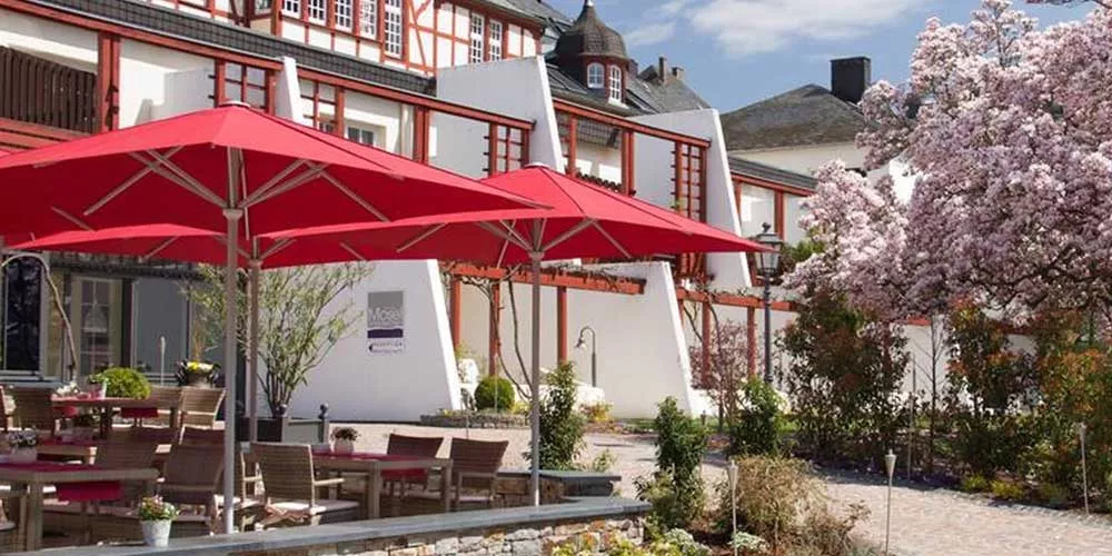 Rote May Sonnenschirme Filius FK auf einer Terrasse vor einem historischen Gebäude mit blühendem Baum im Hintergrund.