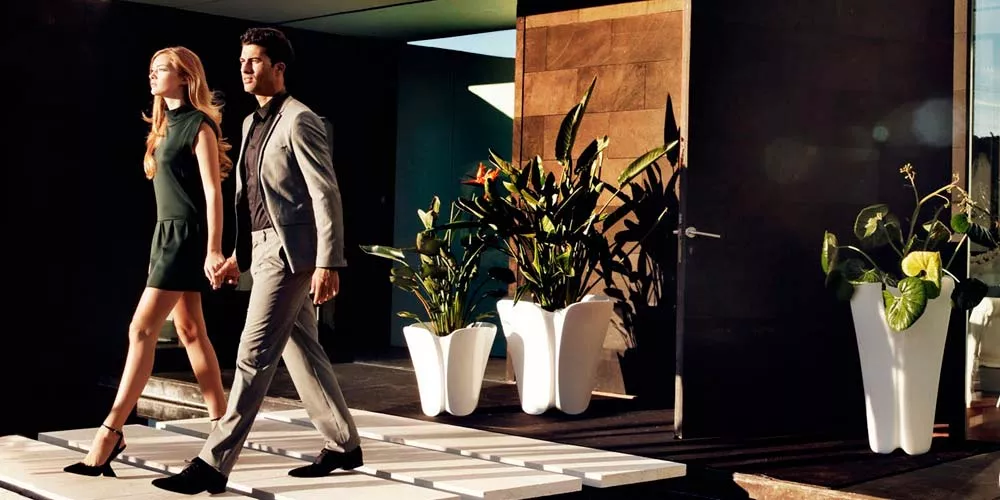 Ein Paar geht an weißen Vondom Pezzettina Pflanzkübeln vorbei, die auf einer modernen Terrasse mit Holzwand im Hintergrund stehen.