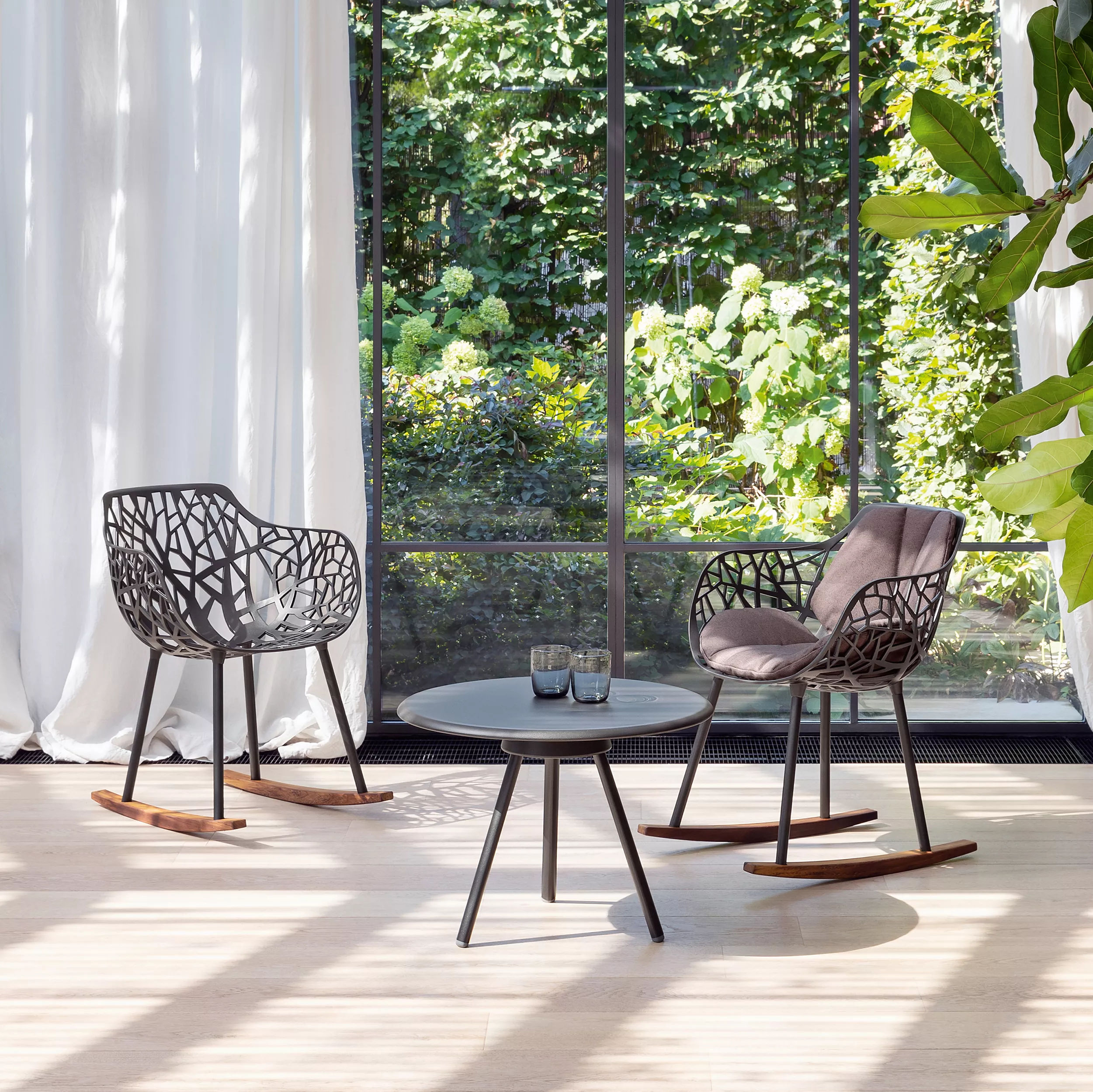 Zwei schwarze Fast Forest Schaukelstühle aus Metall mit Holzbeinen stehen in einem hellen Raum vor einer großen Fensterfront mit Gartenblick.